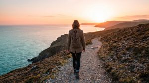 Woman walking a coastal path at golden hour, backpack on, overlooking a calm ocean and distant hills under a vibrant sky.