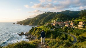 A person views a picturesque Basque village and rugged Cantabrian coastline from a high vantage point at golden hour, with green mountains.