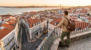 A person with a backpack overlooks Lisbon's red-roofed cityscape and Tagus River with the 25 de Abril Bridge at golden hour.