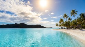Plage de sable blanc avec cocotiers, lagon turquoise limpide et montagnes en arrière-plan sous un soleil brillant.