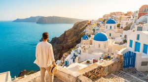 Homme de dos admirant Oia, Santorin : maisons blanches, dômes bleus, mer Égée turquoise et falaises escarpées.