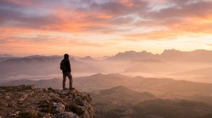 Personne solitaire avec sac à dos sur un sommet rocheux, admirant un vaste paysage montagneux brumeux sous un ciel orangé et rose.