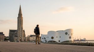 Un homme de dos avec un sac à dos contemple le MuMa et l'église Saint-Joseph du Havre au lever ou coucher du soleil. Un navire est visible au loin.