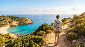 Une randonneuse solitaire sur un chemin côtier surplombant une magnifique crique aux eaux turquoise et une plage de sable.