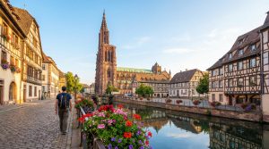 Homme avec sac à dos marche en Petite France, Strasbourg. Canal fleuri, maisons à colombages et la Cathédrale sous ciel clair.