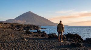 Un homme seul sur une plage volcanique noire contemple un imposant volcan et la mer sous un ciel doux.